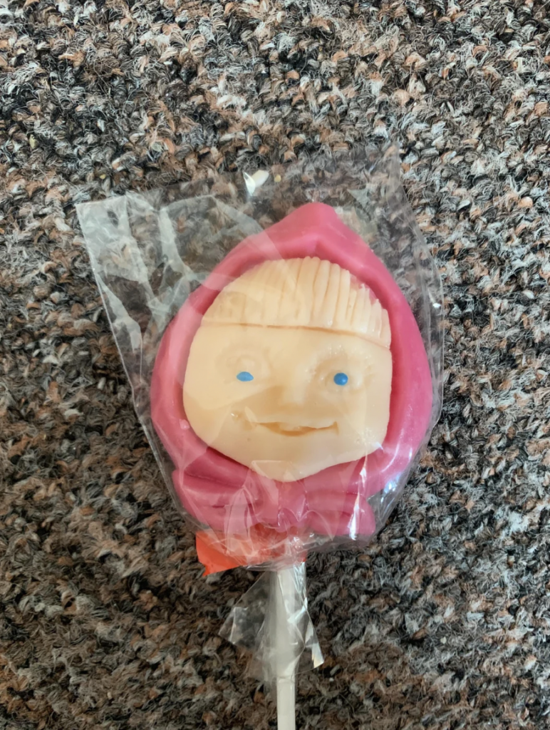 A pink lollipop, shaped and colored like a child's face with blue eyes and a beige face, is wrapped in clear plastic. The lollipop has a hood-like design, resting on a mottled gray and brown carpet background.