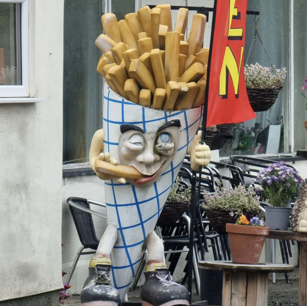 A statue shaped like a cone of fries with a face, arms, and legs stands outside a building. The fries are yellow, the cone has a blue checkered pattern, and the statue is holding a French fry to its mouth. There's a red flag with yellow text and flowers nearby.