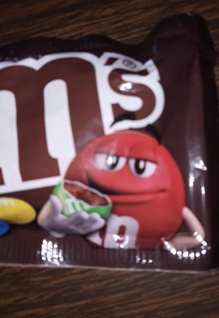 A blurry image of a packet of M&M's featuring the red M&M character holding an opened green M&M's packet, standing next to the large, partial "M" of the M&M's logo. The background is a dark, textured surface.