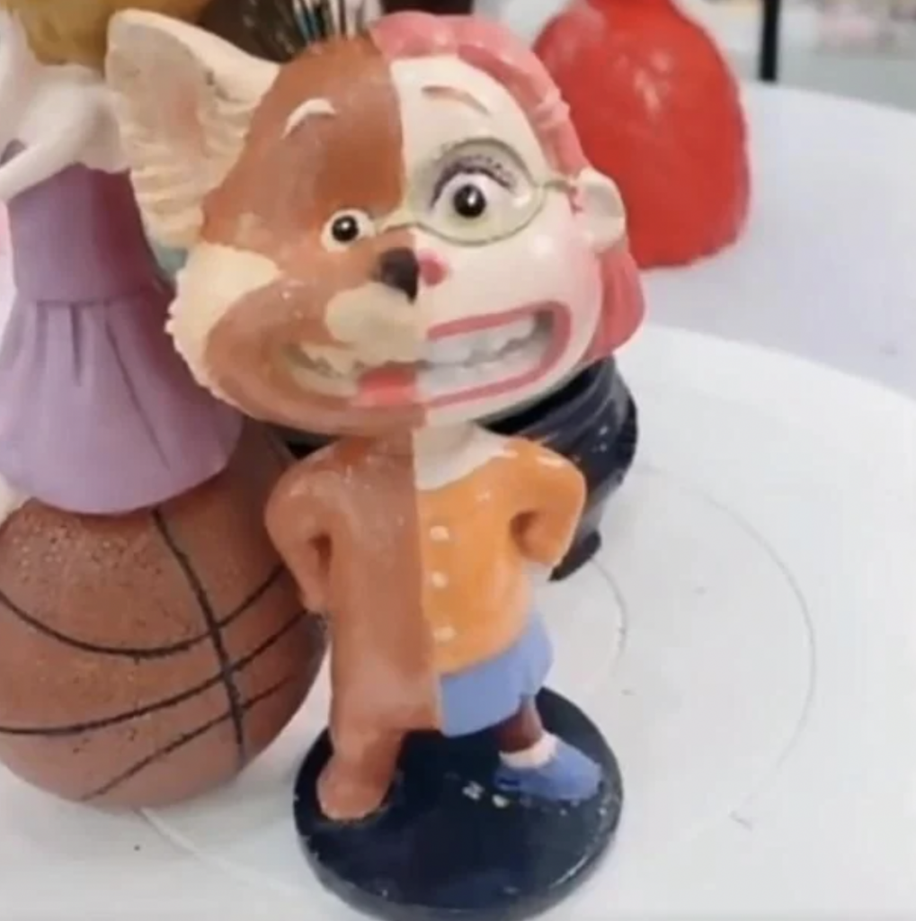 A figurine featuring a character with half of their face resembling a smiling cartoon animal and the other half depicting a smiling human with glasses, wearing an orange top, blue skirt, and blue shoes. A basketball and other figures are partially visible in the background.