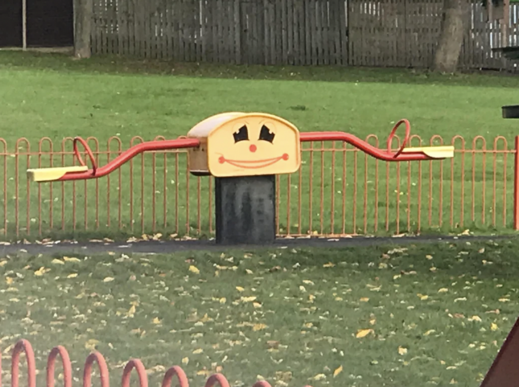 A colorful seesaw with a central structure featuring a smiling face is placed in a grassy playground area. The seesaw has bright red handles and yellow seats, and is surrounded by an orange fence. Leaves are scattered across the grass.