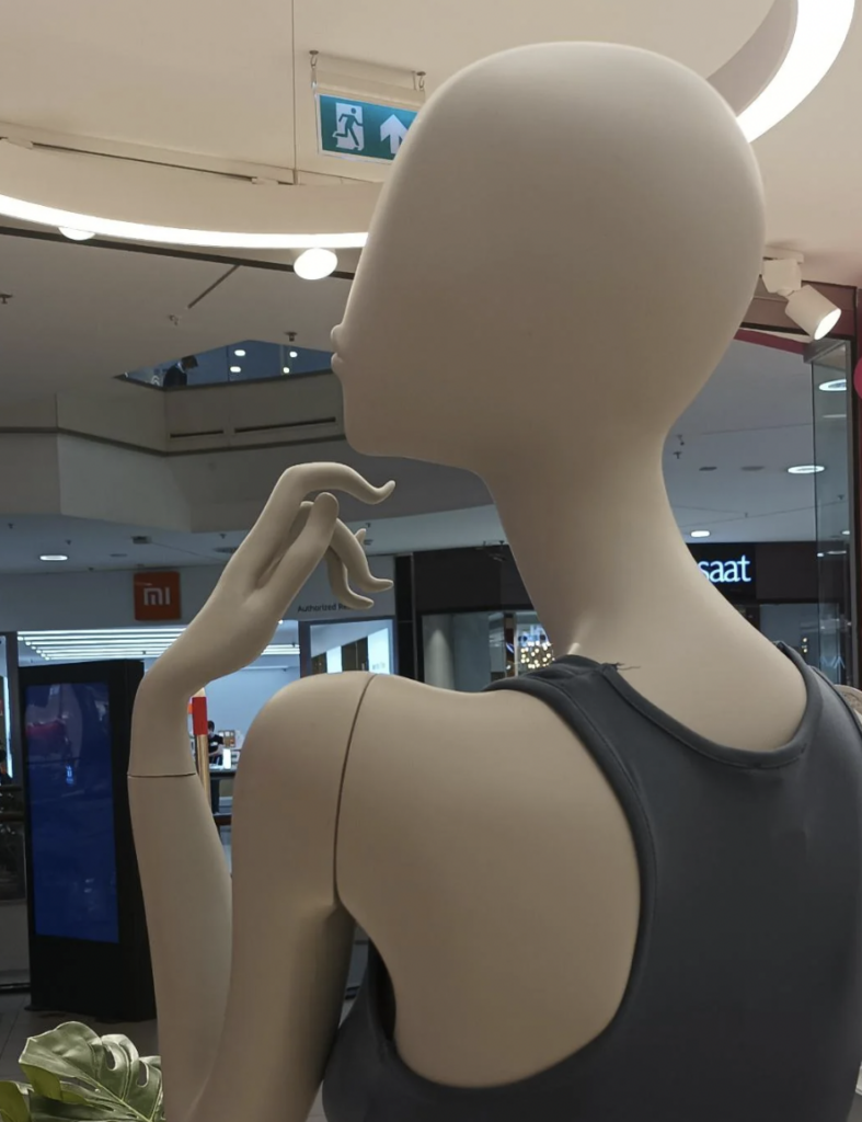 A mannequin dressed in a sleeveless top stands in a shopping mall. The mannequin is posed with a raised arm and fingers near its chin, appearing thoughtful. The background shows various store signs and mall decor.