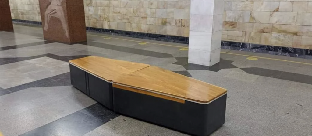 A bench designed to resemble a coffin sits on a tiled floor near a marble wall with decorative patterns. The bench has a wooden lid and dark sides, blending contemporary and gothic aesthetics in a seemingly underground or subway setting.