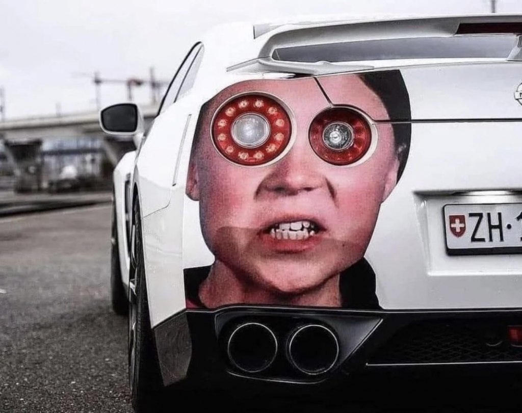 A white sports car with a custom paint job features the face of a person with exaggerated, red circular eyes integrated with the car's rear lights. The license plate has a Swiss flag symbol and the letters "ZH." The background shows an urban area with structures.