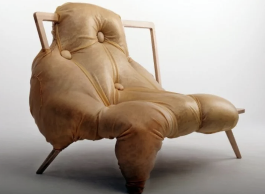 A uniquely designed chair featuring a large, plush, and overstuffed leather cushion with button tufting and a wooden frame. The cushion appears to be sagging comfortably, giving the chair a relaxed and inviting look. The background is a plain, light color.