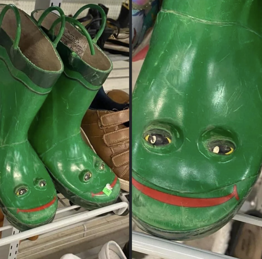 A pair of green children's rain boots with smiling frog faces on the toes. The boots are displayed on a metal shelf, with some other shoes, including a pair of brown ones, visible in the background. The frog faces have eyes and red smiling mouths.