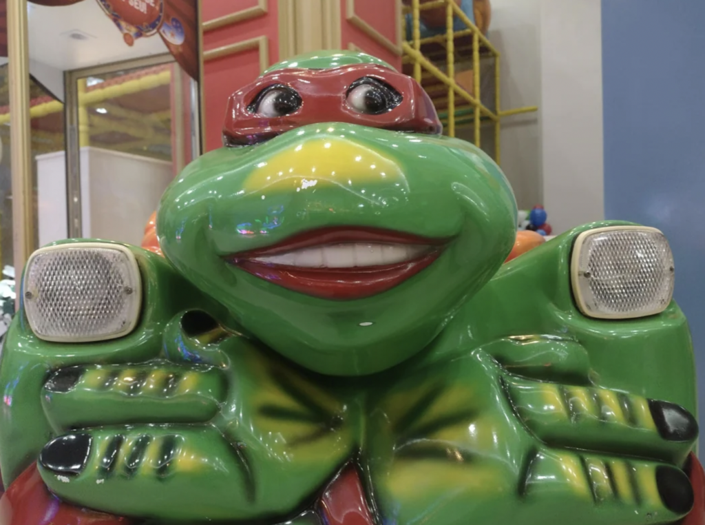 A close-up photo of an amusement ride featuring a colorful character resembling a green anthropomorphic turtle with a red mask. The turtle has a wide, toothy smile and big, round eyes. The ride includes two white lights positioned on either side of its face.