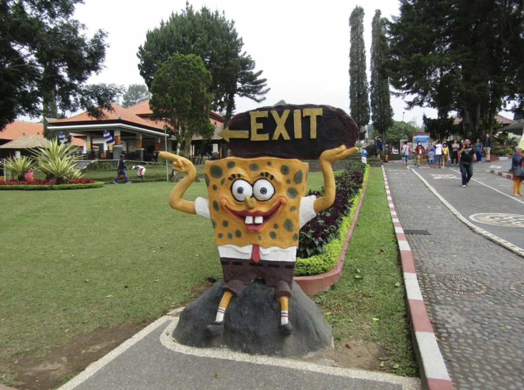 A statue of SpongeBob SquarePants with exaggerated, playful features and his arms raised sits beside a pathway. Above him, a sign reads "EXIT". The scene is set in a park with trees, grass, and paved walking paths. People are visible in the background.