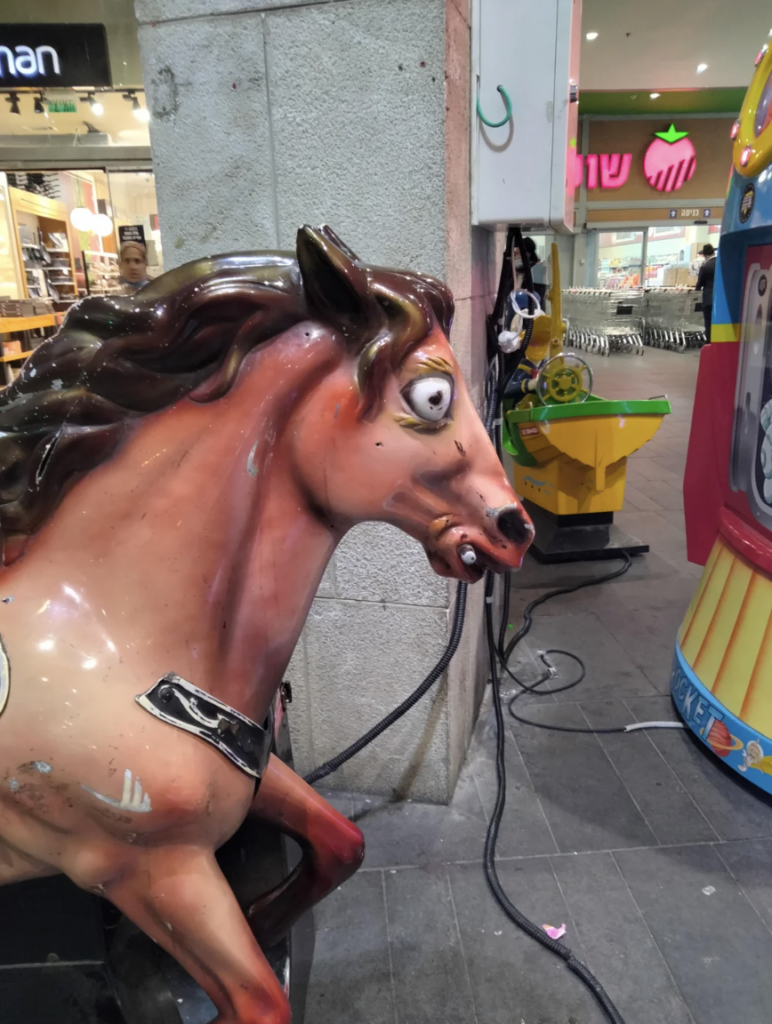 A worn-out, coin-operated horse ride for children is sitting outside a store in a shopping mall. The horse has a scruffy side and looks toward the ground. Some other kids' rides and shopping carts are visible in the background.