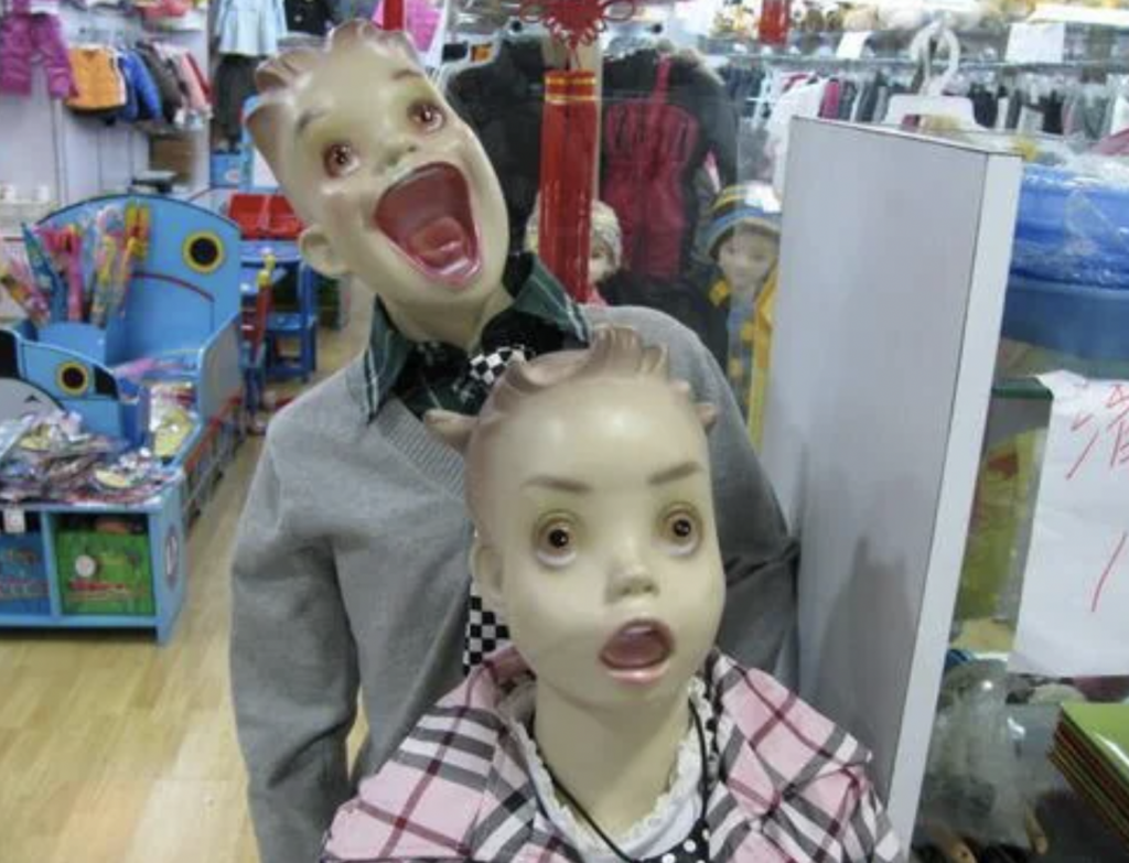 Two mannequins with exaggerated, surprised expressions stand inside what appears to be a store. The background includes shelves with various items and colorful decor. The front mannequin is wearing a plaid jacket, while the one behind wears a grey sweater.