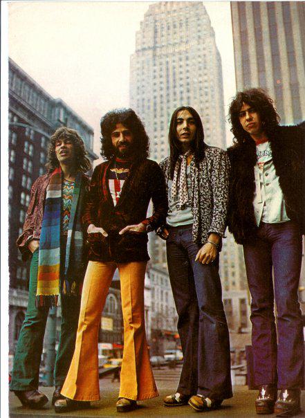 Four men stand on a city street in front of tall buildings. They are dressed in colorful 1970s attire, including flared pants, scarves, and jackets. The background shows a bustling urban environment with cars and pedestrians.