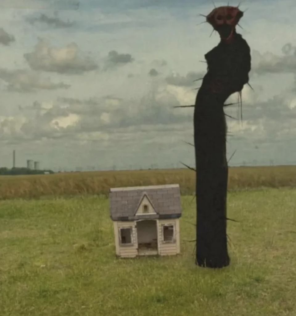 A surreal scene showing a tiny, white, single-story house with a gabled roof on a grassy field. Next to the house is a towering, black, spiky, humanoid figure with glowing red eyes, casting a shadow. The background has a cloudy sky and distant industrial buildings.