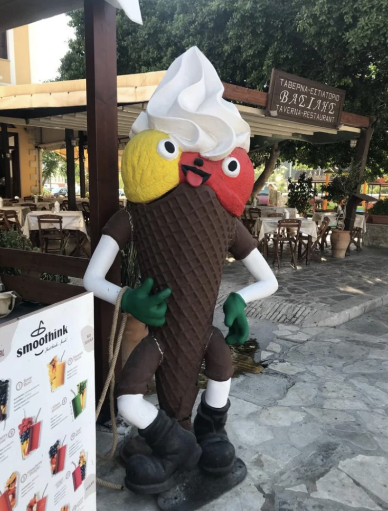 A whimsical statue of an ice cream cone with a waffle cone body, legs, and arms. The cone has scoops of yellow and red ice cream with a swirl of white on top, big googly eyes, and a wide smile. It stands next to a menu stand for "Smoothik" at an outdoor restaurant.