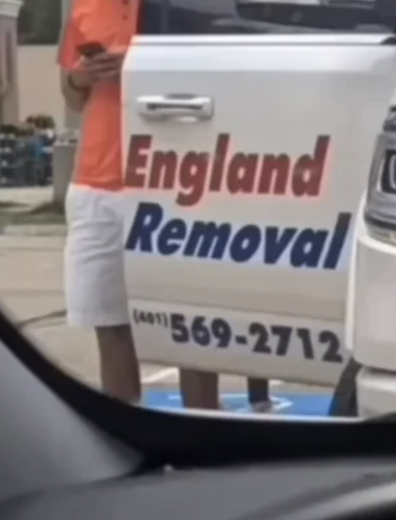 A person in a bright orange shirt and white shorts stands next to a white vehicle with the words "England Removal" and a phone number written on the door. The image is taken from inside another car, partially showing the dashboard and window frame.