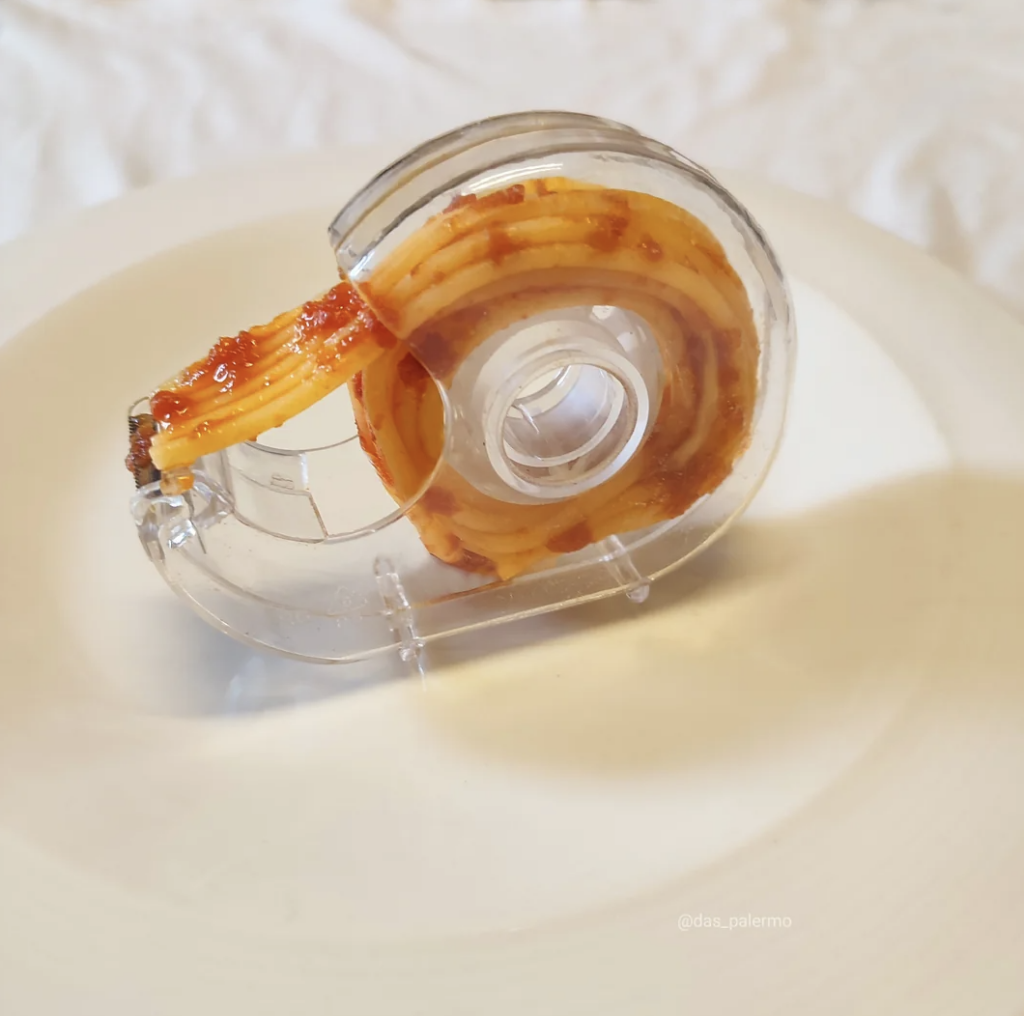 A transparent tape dispenser is filled with a coil of spaghetti instead of tape. The spaghetti appears to have a tomato-based sauce on it. The dispenser is placed on a white surface.