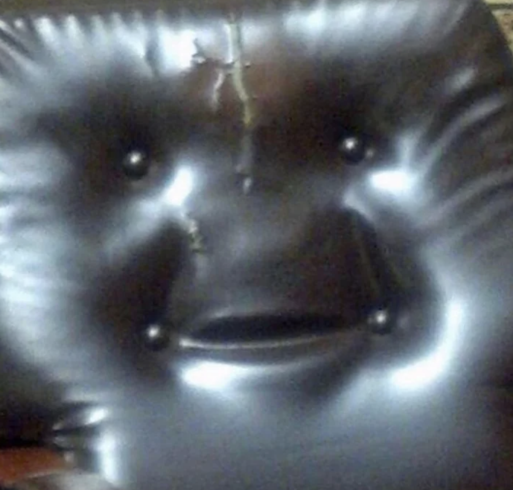 A close-up photo of a worn-out black leather cushion. The stitching and indents on the cushion resemble a smiling face with two small buttons for eyes, a slit for a mouth, and a thin vertical tear on the forehead.