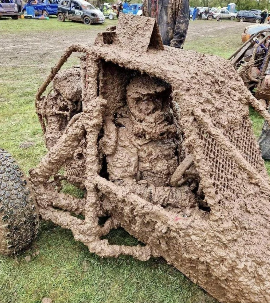 A heavily mud-covered off-road vehicle with a driver inside, equally plastered with mud, sits on a grassy area. The vehicle's frame, wheels, and protective netting are barely visible underneath the thick layer of mud. Other vehicles are seen in the background.