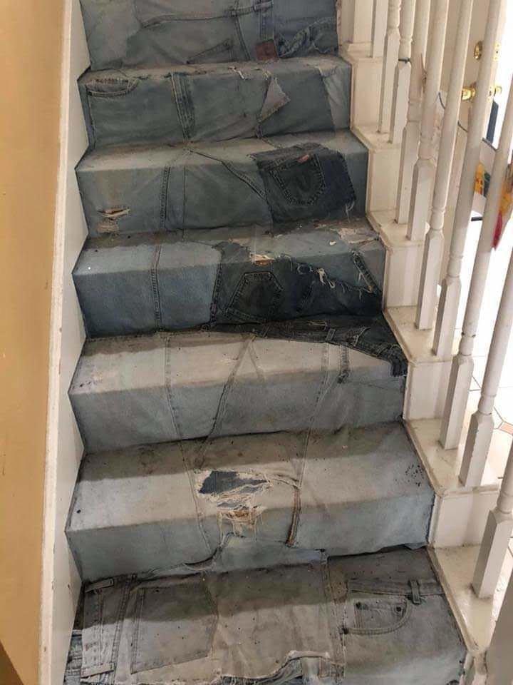 A staircase is covered with worn and torn blue jeans. Each step is wrapped in a different pair of jeans, showcasing various shades of denim and patterns of wear and tear. The handrail on the right side of the staircase is white.