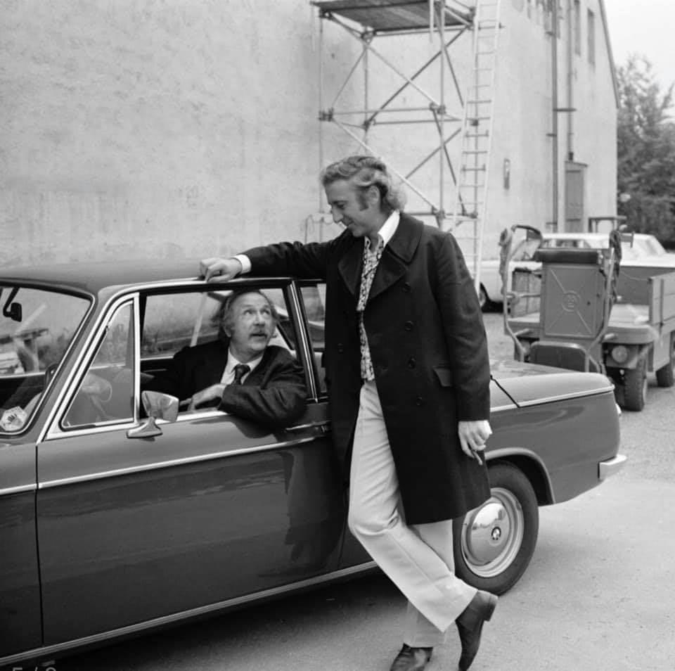 Two men are conversing beside a parked car. One man is seated inside the car, leaning out of the window, engaged in conversation with the other man who is standing and leaning on the car roof. There is scaffolding and construction equipment in the background.