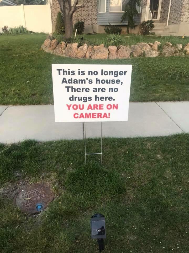 A sign in a residential yard reads, "This is no longer Adam's house. There are no drugs here. YOU ARE ON CAMERA!" The sign is placed near a sidewalk, a patch of grass, and a small utility cover. A house and landscaping are visible in the background.