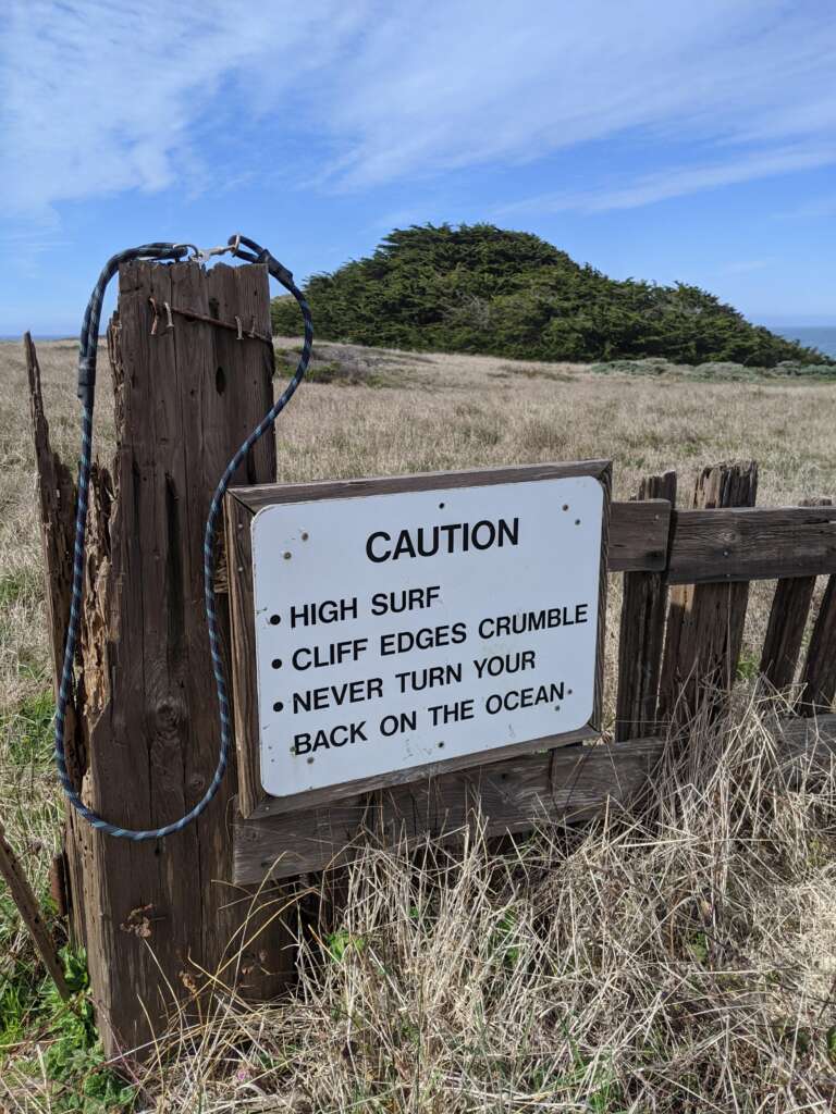 A weathered wooden post and fence with a sign that reads, "CAUTION: High surf, Cliff edges crumble, Never turn your back on the ocean." The fence is overlooking a grassy field with a hill and trees in the distance under a partly cloudy sky.