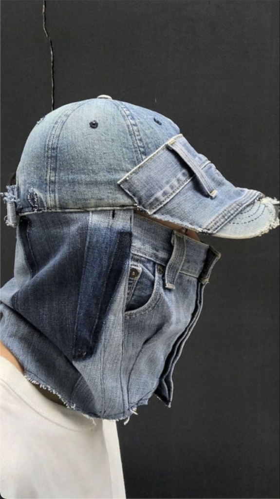 A person wearing a unique hat made from repurposed denim jeans, with pockets and seams clearly visible. The hat has a visor and an extended section that covers the lower half of the face, resembling a mask. The backdrop is black.