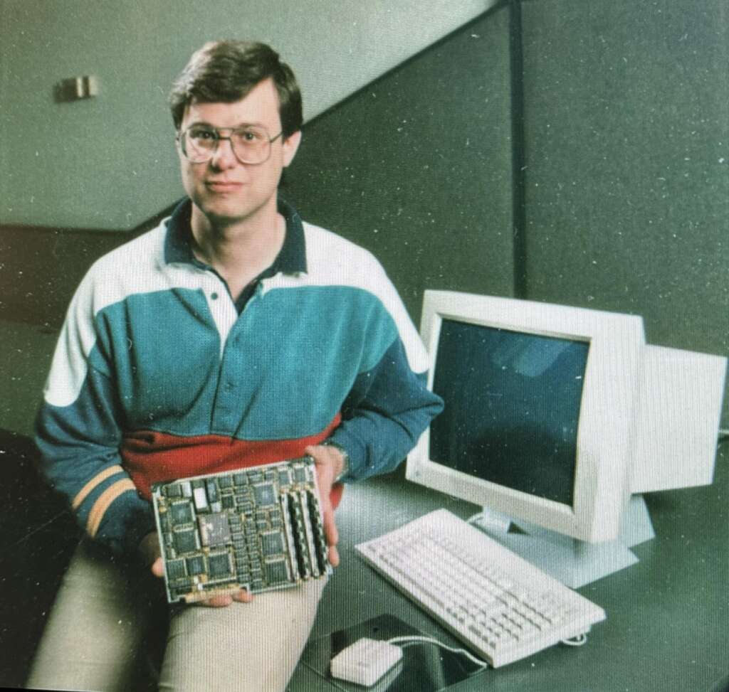 A man wearing glasses and a color-blocked sweatshirt holds a computer motherboard. He is sitting next to an old desktop computer setup, which includes a CRT monitor, keyboard, and a mouse, all placed on a desk. The background suggests an indoor office environment.