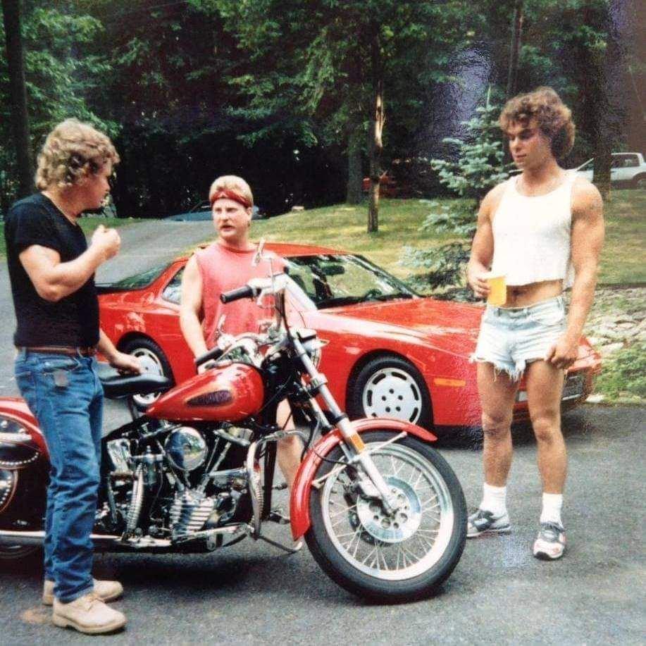 Three men stand outside near a red motorcycle and a red sports car. One man on the left is wearing a black shirt and jeans, the middle man is wearing a red sleeveless shirt, and the man on the right is in a white crop top and ripped denim shorts, holding a yellow mug.