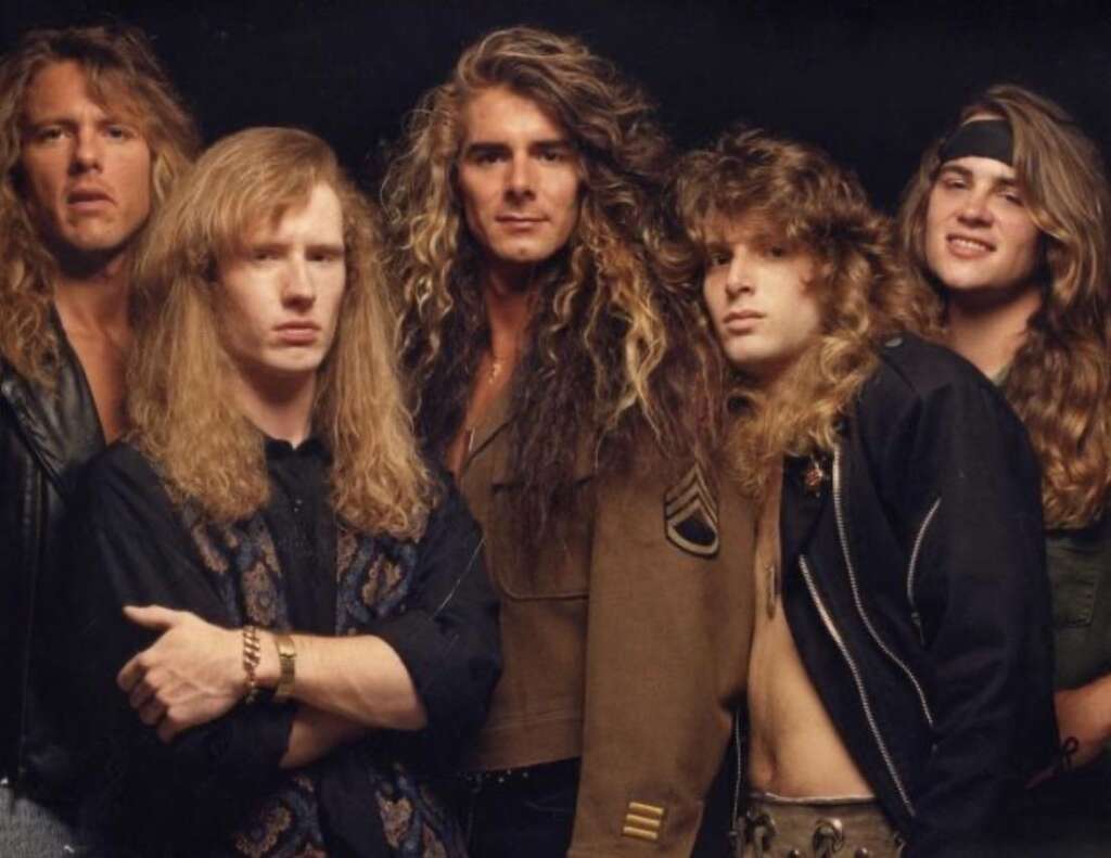 Five men with long hair and 80s rock style clothing pose together against a dark backdrop. They wear leather jackets, vests, and casual shirts. Some have serious expressions while others smile casually.