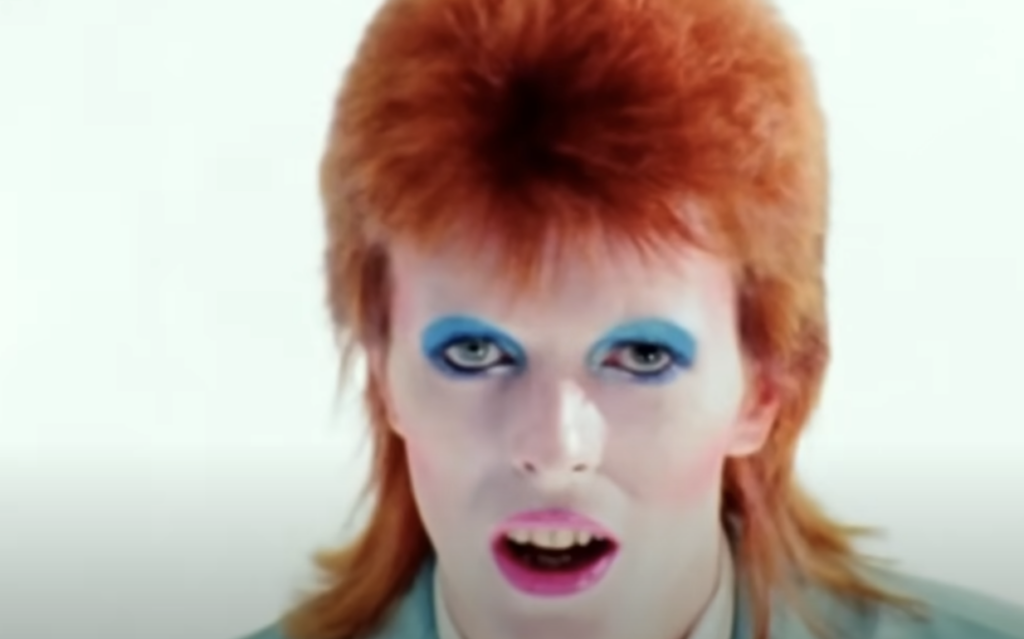 A person with bright orange hair is wearing heavy blue eyeshadow and dramatic makeup. They have a pale face and are looking directly at the camera with their mouth slightly open. The background is white.