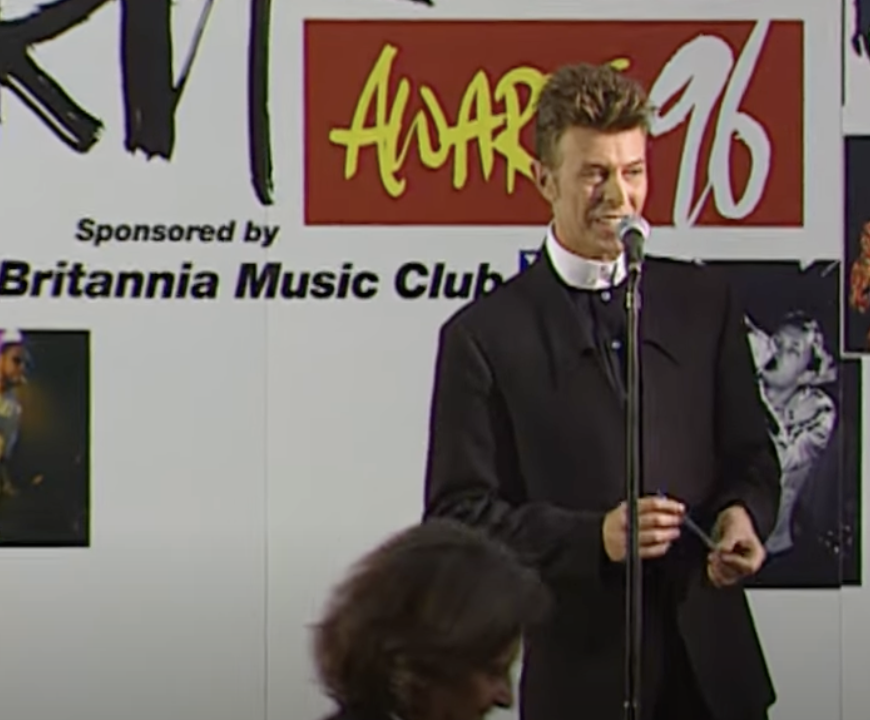 A person in a dark suit stands smiling at a microphone on a stage, against a backdrop featuring the text "Awards 96" and "Britannia Music Club." Other figures and images are partially visible in the background.