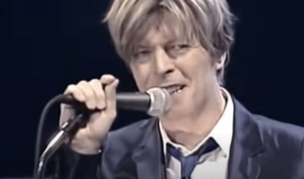 A person with light brown hair is singing into a microphone. They are wearing a dark suit with a white shirt and loosely tied blue tie. The background is dark, suggesting a stage or performance setting. The person appears to be smiling or in mid-speech.