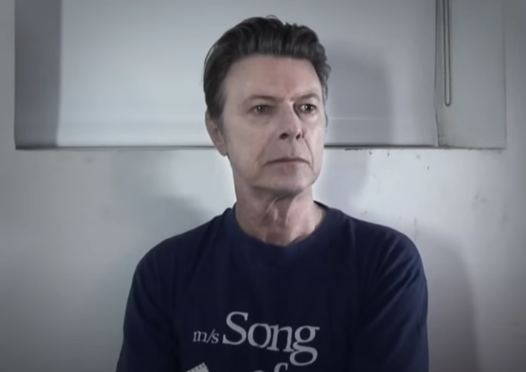 A person with short, styled hair is standing in front of a white background, wearing a navy blue shirt with partially visible white text that reads "Song." The individual appears to be looking slightly to the left with a neutral expression.