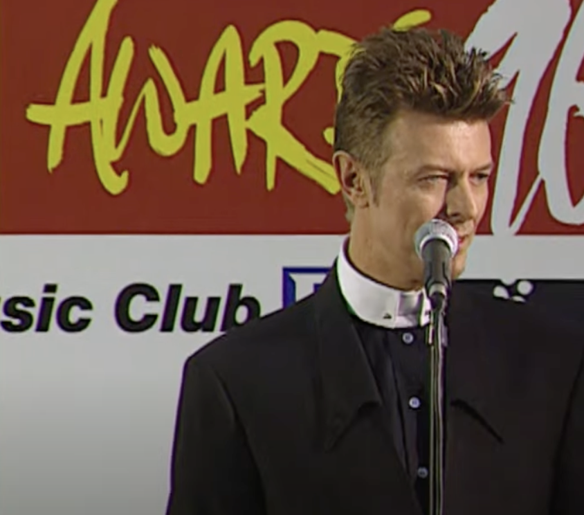 A man stands at a microphone, dressed in a dark suit with a white collar, against a backdrop featuring the word "Awards" in yellow and red text, and the phrase "Music Club" partially visible. He has styled brown hair and is looking to the side.