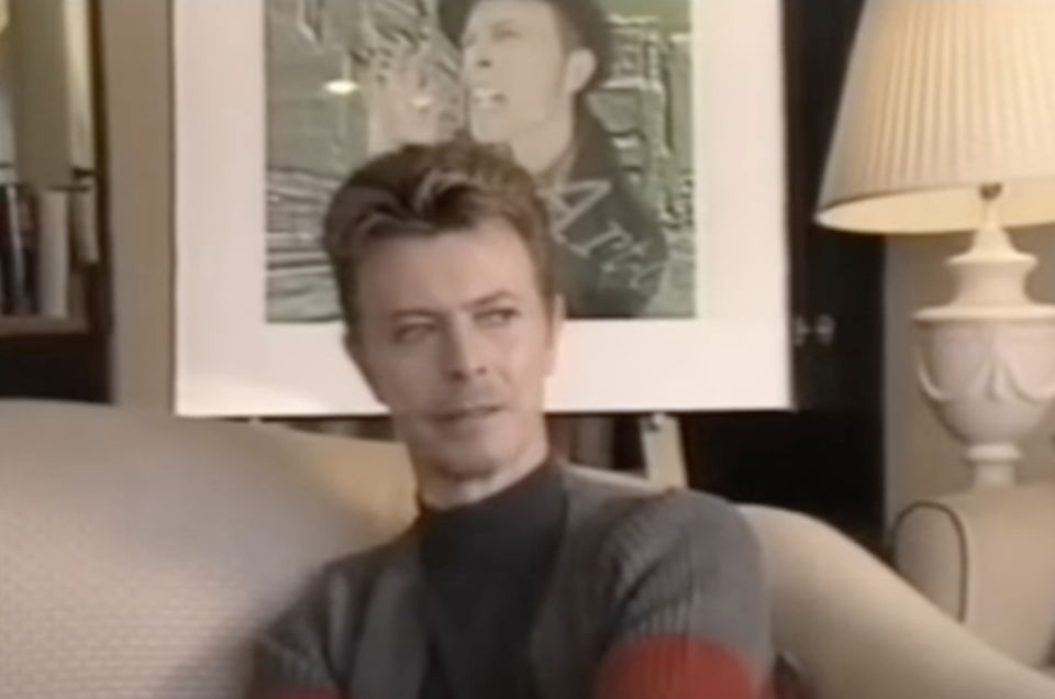A man with short, styled hair is sitting on a sofa. He is wearing a dark turtleneck sweater with red accents on the sleeves. Behind him, there is a framed photo of another individual singing passionately, displayed on the wall. A lit table lamp is also visible.