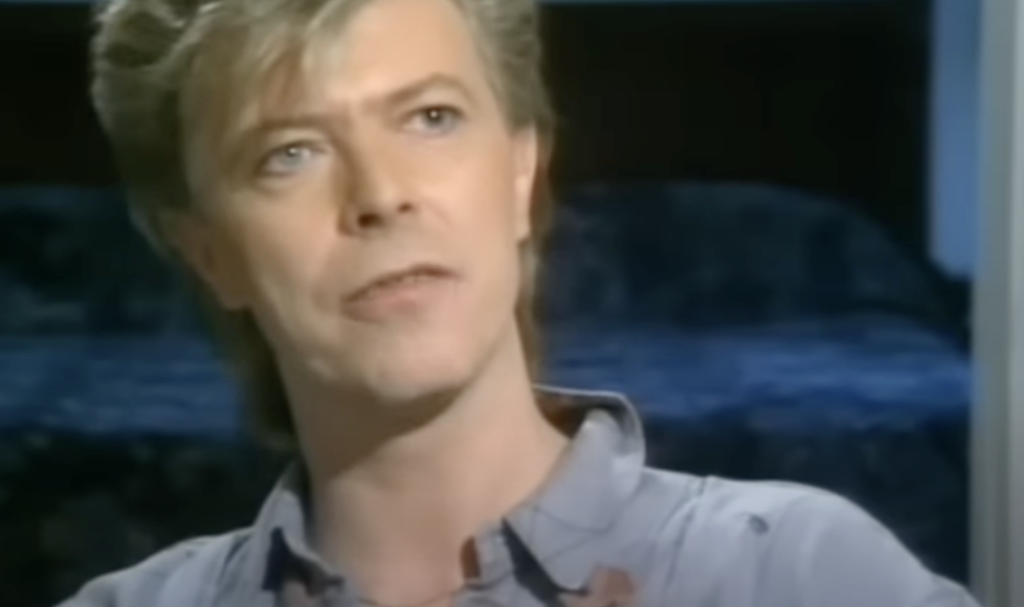 A person with short, light-colored hair is looking slightly to the side while speaking. They are wearing a light-colored button-up shirt and are in front of a blurry, dark background.