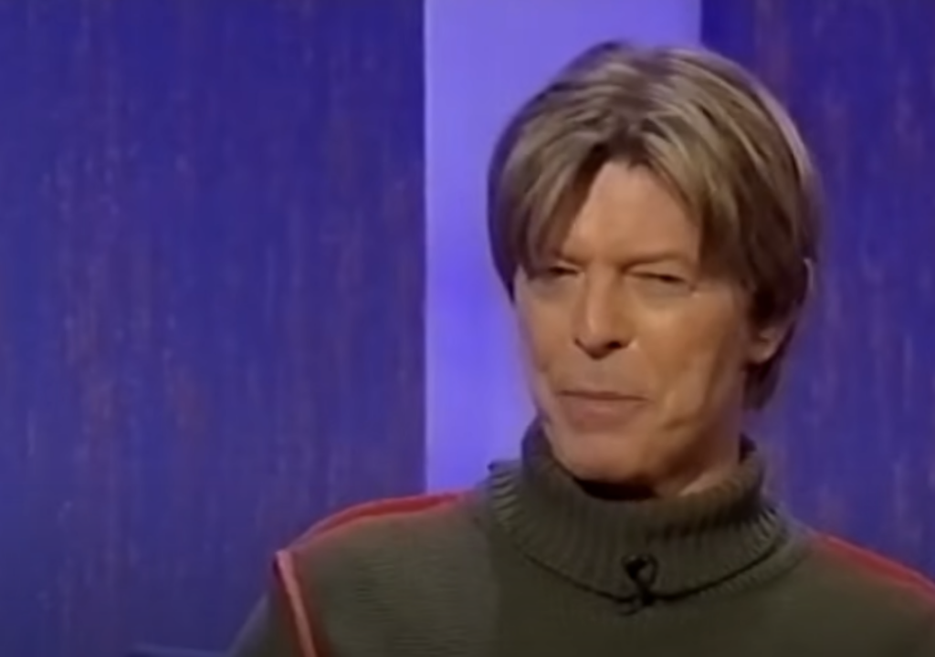 A person with short, light brown hair is sitting indoors against a purple and blue background. They are wearing a grey, high-neck sweater with a red stripe. The person appears to be in mid-conversation or making a speaking expression.