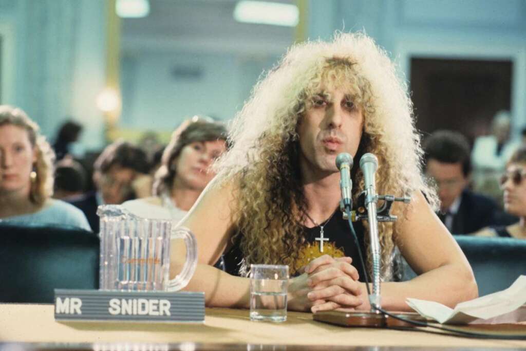 A person with long, curly blonde hair and a black sleeveless top speaks into a microphone at a table labeled "Mr. Snider." There is a water pitcher and glass on the table. The background shows an audience seated and listening attentively.