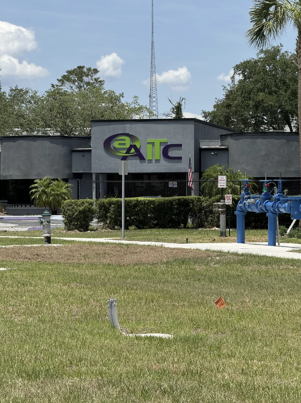 A gray building with "EaTC" logo on the front is surrounded by greenery, including a lawn and hedges. A blue fire hydrant is visible on the right side, and an American flag is positioned near the entrance. The background shows trees and a partly cloudy sky.