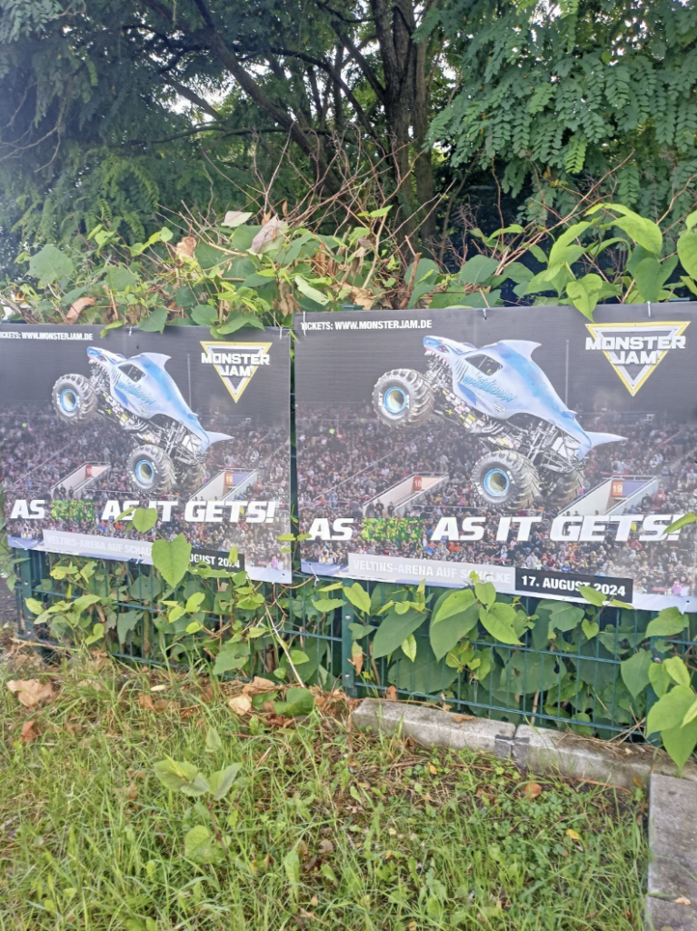 Posters for "Monster Jam: As It Gets" are displayed outdoors on a fence, depicting a blue monster truck in mid-air with a backdrop of a cheering crowd. The event is scheduled for August 17, 2024, at the Getec Arena. Green foliage partially covers the posters.