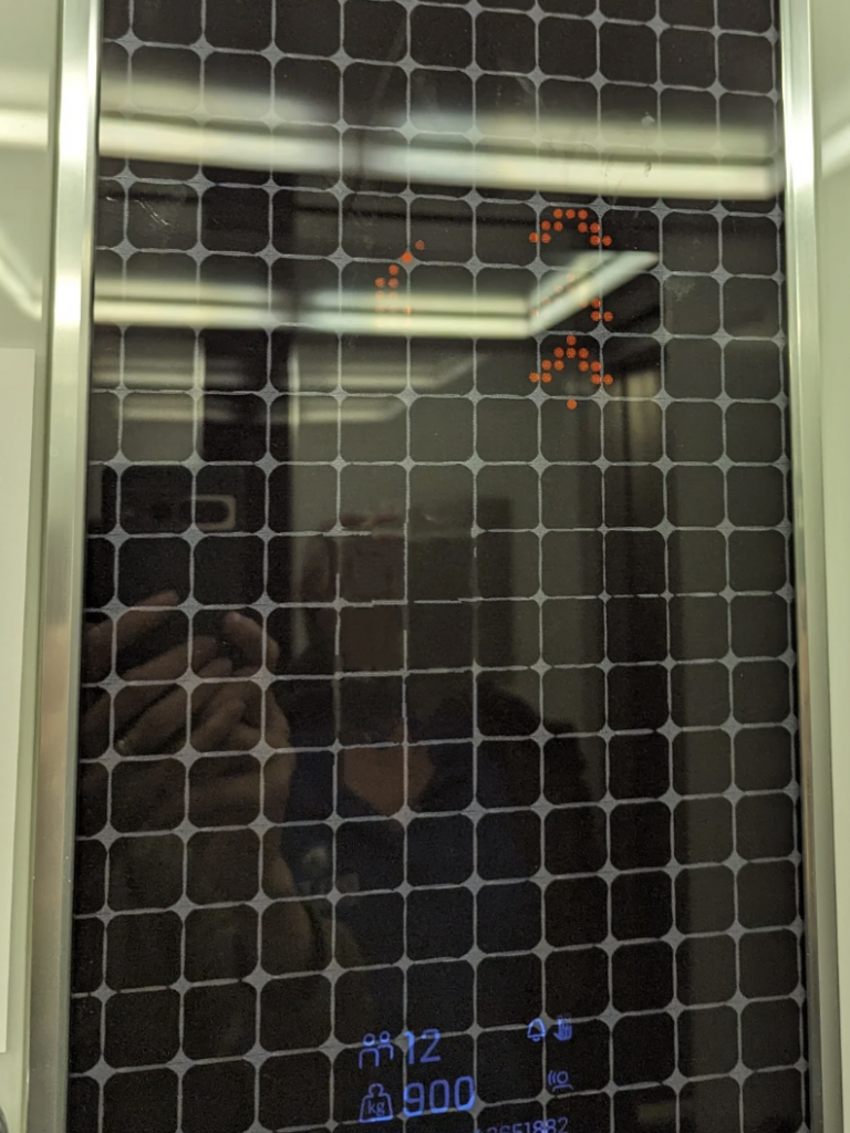A digital elevator display shows the elevator is on the 12th floor with a maximum capacity of 900 kilograms. Red LED arrow indicators are pointing upwards. A faint reflection of a person taking the photo is visible on the screen.