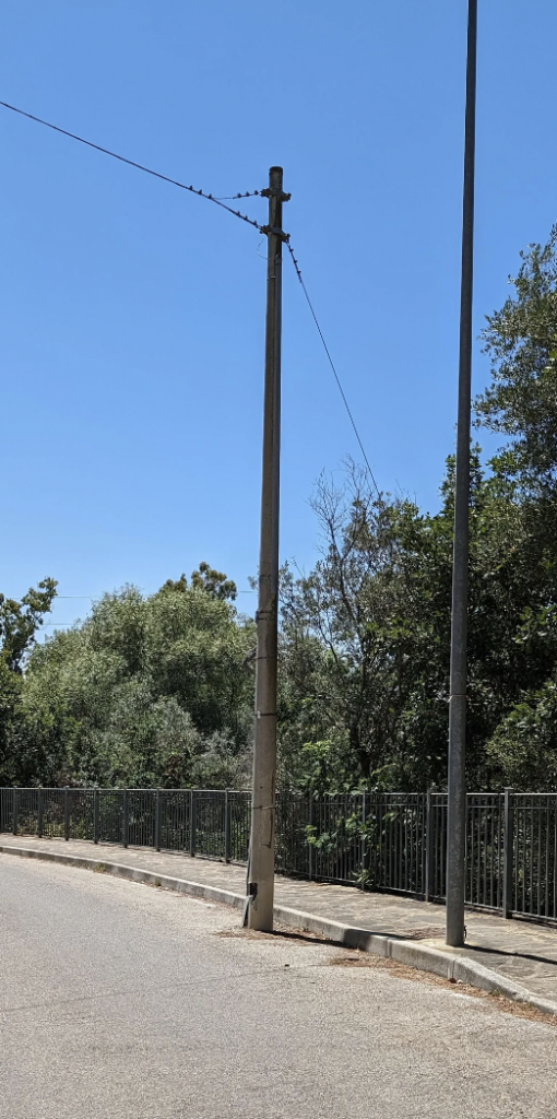 A street scene featuring a tall concrete utility pole with cables attached, standing near a road. To the right of the utility pole is a lamppost. In the background, there are lush green trees and a metal fence running alongside the road. The sky is clear and blue.
