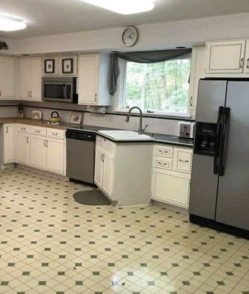 A spacious kitchen features white cabinets, a stainless steel dishwasher, and a matching side-by-side refrigerator. The tiled floor has a light green pattern. A large window above the sink lets in natural light, and a clock is mounted above the cabinets.