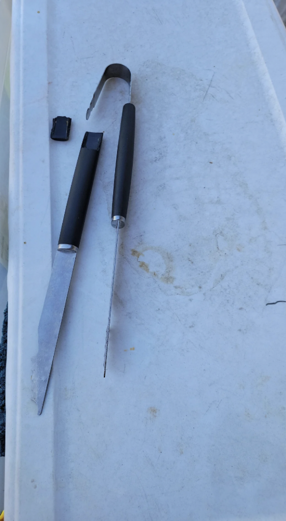 A broken knife with a detached blade and handle lies on a light gray surface. The handle has a black grip, and the blade is thin and metal. Nearby, a detached plastic piece and what appears to be a metal clip are also visible.