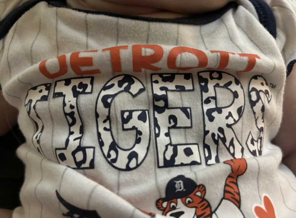 Close-up of a baby's white onesie featuring "Detroit Tigers" text. The letters in "Tigers" have a tiger stripe pattern. Below the text, a cartoon tiger wearing a Detroit baseball cap is depicted holding a bat.