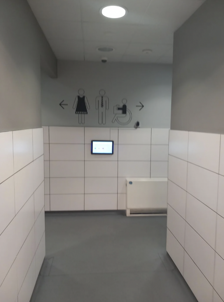 A hallway with white tiled walls leads to a gender-inclusive and accessible restroom. The wall at the end of the hallway has icons indicating a men's, women's, and wheelchair-accessible restroom. A touchscreen panel is mounted below the icons.