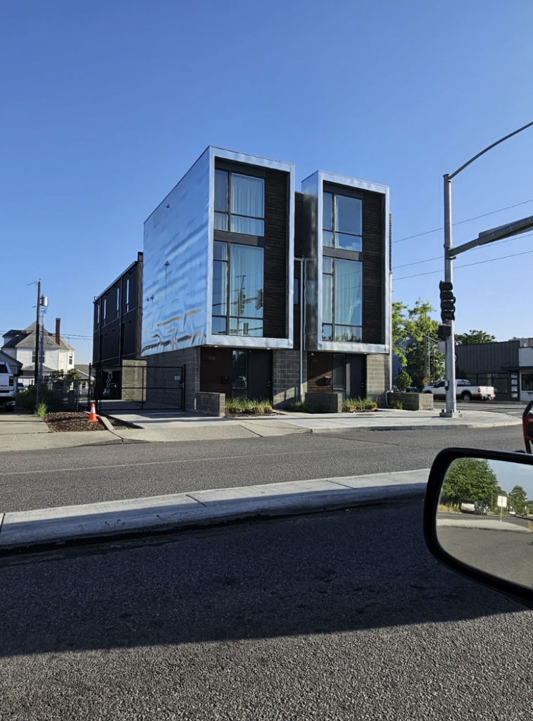 Modern, minimalist building with two adjacent rectangular structures featuring large windows and a metallic facade. The building is situated next to a street with a traffic light in view and clear skies above. A side mirror of a car is visible on the right edge of the image.