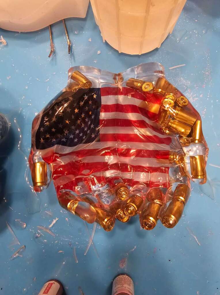 A clear resin sculpture of two cupped hands holding bullet casings, with an American flag embedded in the palms. The sculpture is placed on a light blue surface with a few tools and materials surrounding it.