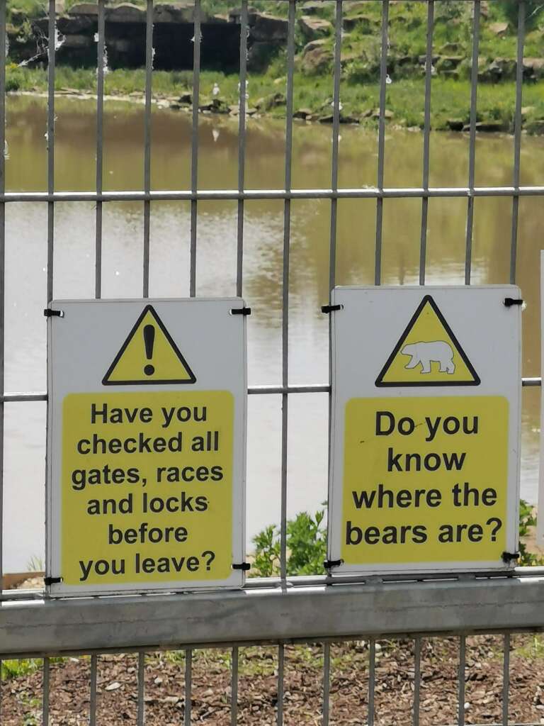 Two warning signs on a metal fence in front of a body of water with trees and rocks in the background. The sign on the left asks, "Have you checked all gates, races and locks before you leave?" The sign on the right asks, "Do you know where the bears are?