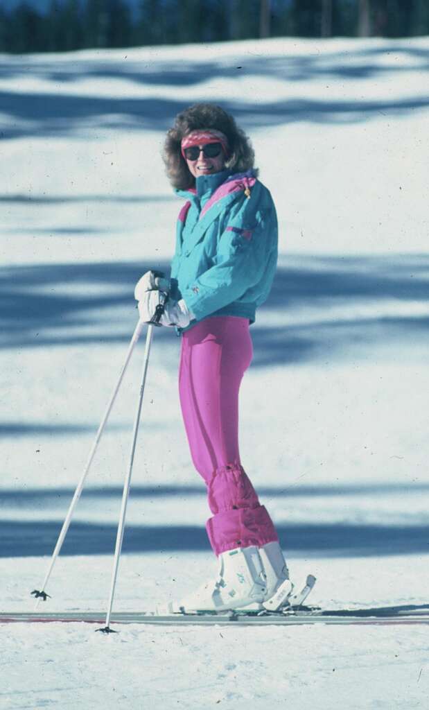 A person stands on a snowy slope wearing a turquoise jacket, pink ski pants, and pink goggles, holding ski poles. They are wearing white ski boots and have a hood lined with fur. The background is a snowy landscape with trees faintly visible in the distance.